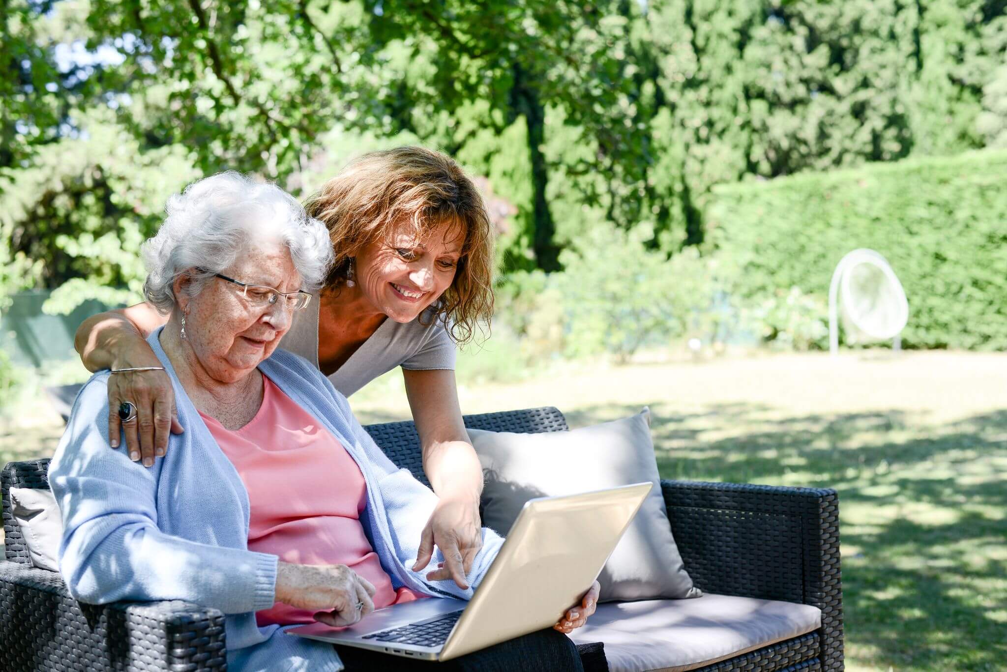 A woman site outside in a couch with a friend who is helping her with a computer