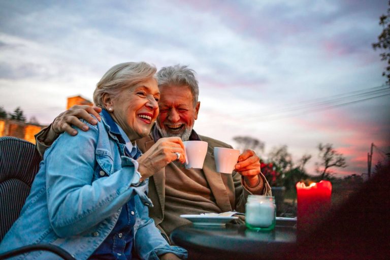 A couple sits at an oudoor table and hold up mugs of coffee
