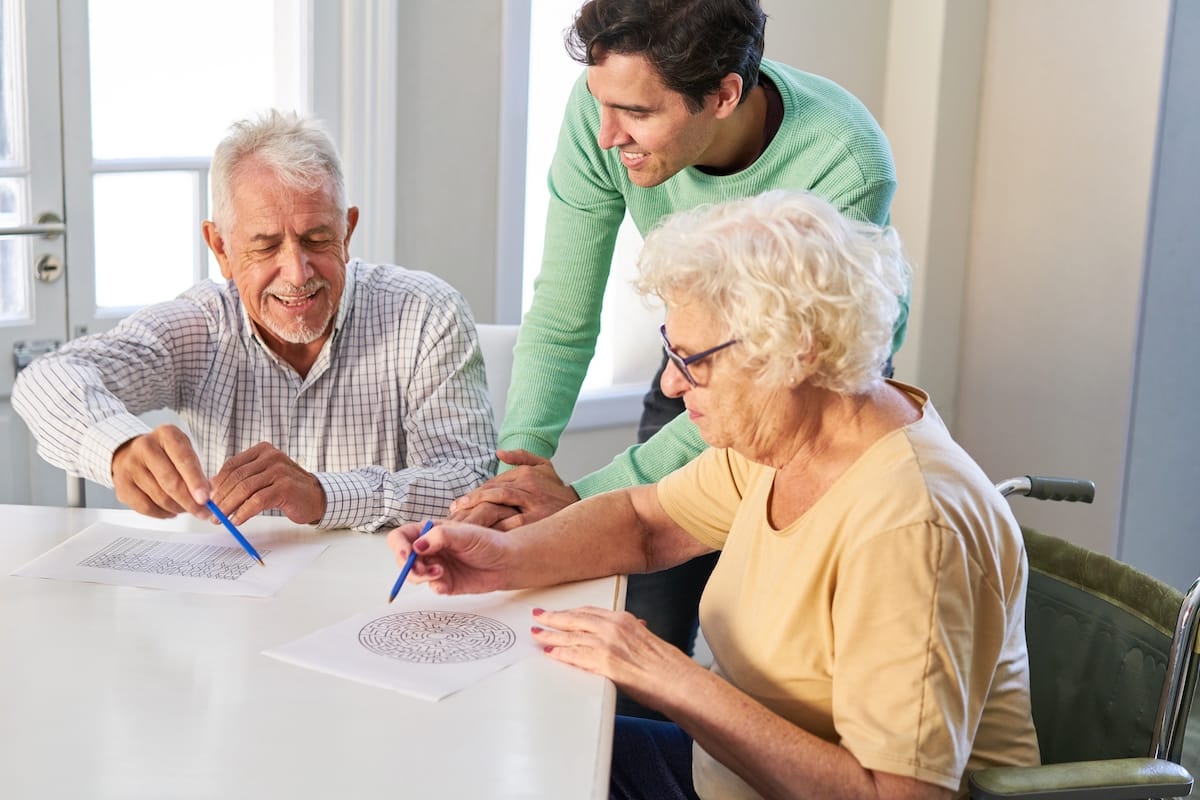 SeniorCoupleWithSonSolvingMazePuzzlesAsMemoryTraining - Rockbridge Oaks Caregiver helping older adults with a puzzle